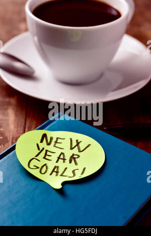 a yellow sticky note in the shape of a speech balloon with the text new year goals written in it attached to a black notepad on a rustic wooden table Stock Photo