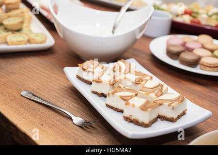 Vanilla Cheesecake squares swirled with caramel, with cold cuts and quiche appetizers in the background Stock Photo