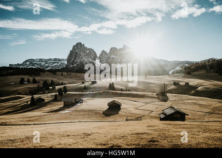 Wooden houses on a field at the foot of a mountain range. Photographed at sunrise with the sun as a backlight. Stock Photo