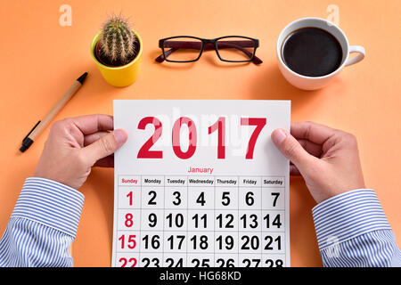 a young caucasian businessman observes a 2017 calendar sitting at his office desk, where there is a pen, a cactus, a pair of eyeglasses and a cup of c Stock Photo