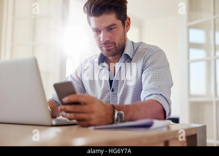 Shot of confident young business man reading text message on his mobile phone while working on laptop. Young man working from home office. Stock Photo
