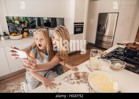 Happy young mother taking selfie with her daughter in kitchen. Young woman and little girl making food in kitchen taking self portrait with mobile pho Stock Photo