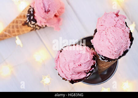 Top view of pink strawberry ice cream in waffle cones shot over a rustic wooden background with fairy lights and more icecream. Extreme shallow depth Stock Photo