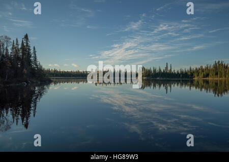 Dead calm lake in early morning with reflections of conifers and wispy cirrus clouds Stock Photo