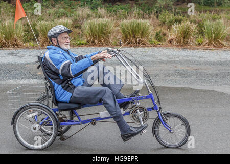 A smiling, elderly man, age 94, pedals a recumbent tricycle along a driveway, equipped with a helmet, flag and other safety gear. Stock Photo