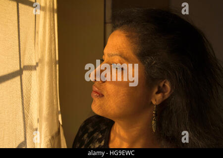 Young Indian woman looking at window with golden light on her face Stock Photo