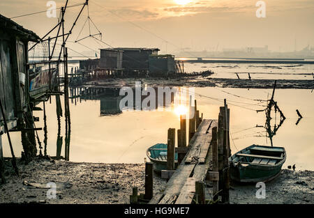 Old fishing huts reflecting in the lagoon of Marina di Ravenna at sunset, industrial harbor on background. Stock Photo
