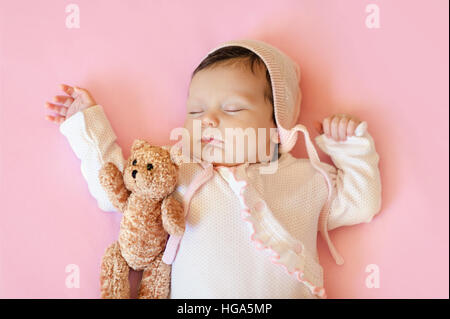 baby girl in white hat and pink pajamas hugging teddy bear sleeps in his bloodstream on a  blanket Stock Photo