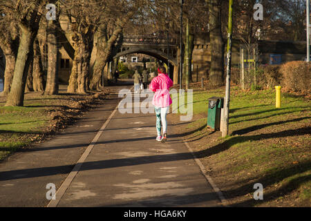 Female jogger running along tree-lined, riverside, footpath towards Skeldergate Bridge on a sunny, winter day - York, England.