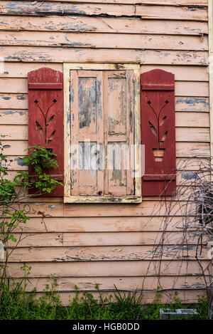 Building wall with pink, tan peeling paint, a shuttered window and red decorative shutters. Stock Photo