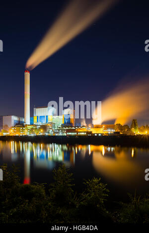 Colorful night shot of an illuminated modern industrial building with steam and deep blue sky behind a canal. Stock Photo