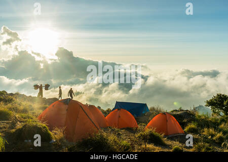 Tent camp on the crater rim of Mount Rinjani, Lombok, Indonesia. Sun is setting in the clouds, tents glow bright orange. Stock Photo