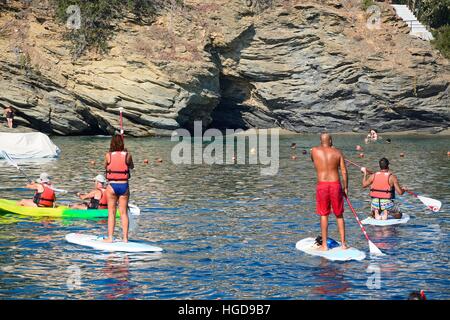 Holidaymakers on paddle boards with a small beach and cave to the rear, Bali, Crete, Greece, Europe. Stock Photo
