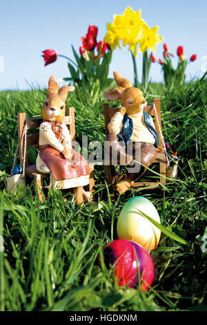 Married Easter bunny couple sitting on chairs in a meadow with daffodils and tulips Stock Photo