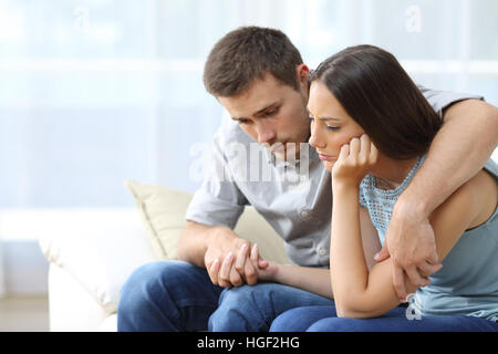 Sad couple comforting each other sitting on a couch in the living room at home Stock Photo