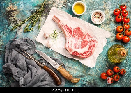 Raw tomahawk beef steak with ingredients for grilling: seasoning, fresh rosemary and olive oil on paper, top view Stock Photo