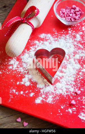 Heart shape cookie cutter, flour and roller over wooden and red background - love, romance, baking with love, valentine day festive homemade pastry co Stock Photo