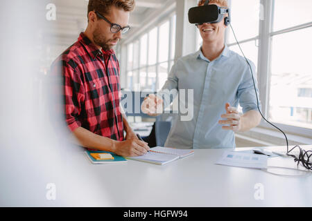 Young man writing notes with colleague wearing virtual reality goggles in office. Developers working on augmented reality device technology. Stock Photo