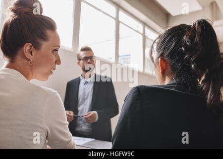Two young woman discussing during in a meeting in office. Businessman standing in background  with female colleagues talking in front. Stock Photo