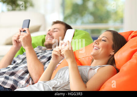 Couple using their smart phones lying on a green and orange poufs in the living room at home Stock Photo