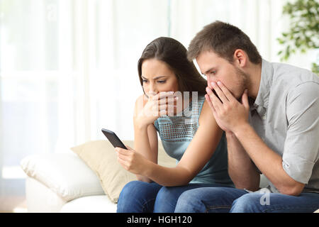 Worried couple reading bad news together on the phone sitting on a sofa in the living room at home Stock Photo