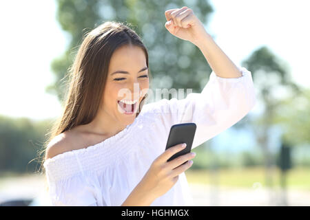 Excited beautiful girl receiving a sms message with good news in a mobile phone outside in a park with a green background Stock Photo