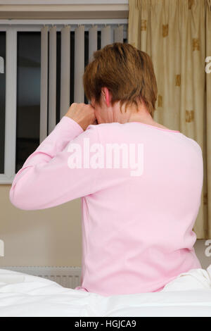 Back view of a tired mature woman rubbing eyes sitting up on a bed awake at night suffering from insomnia due to stress at bedtime. England UK Stock Photo