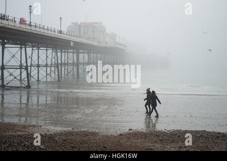 Two people walk along the beach during low tide on the beach in front of Worthing Pier in Sussex, England on a foggy day. Stock Photo