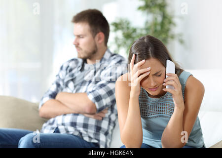 Victim of gender abuse calling on smartphone for help sitting on a couch at home with her husband beside Stock Photo