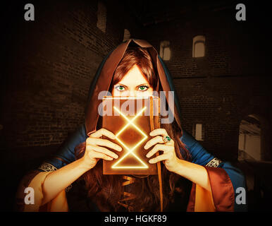 Beautiful young halloween witch wearing vintage gothic dress with hood holding magical book of spells in old leather cover inside ancient dark castle Stock Photo