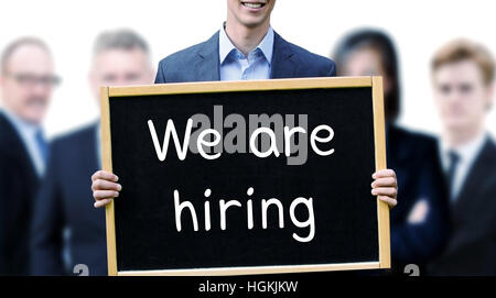 young businessman holding a sign with the words We Are Hiring and group of people behind him Stock Photo