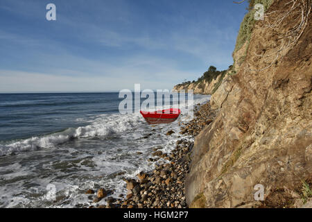 Red rowboat in the surf on a rocky beach beneath a cliff. Stock Photo