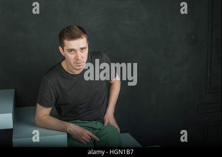 studio photography young brutal guy. man in T-shirt on a background of black wall on the white steps. sitting on the steps, looking at the camera Stock Photo