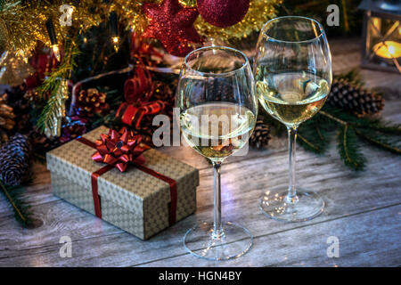 Two wineglas with white wine on a wooden table in the background of Christmas tree, gift and candles Stock Photo