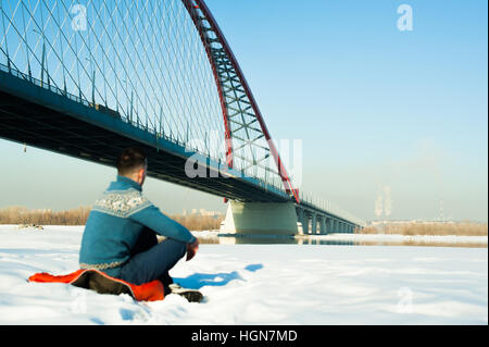 man in warm clothes with a beard is walking on the street in the winter in a warm sunny day at the river bridge in the background, sits in the snow Stock Photo