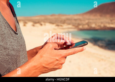 closeup of a young caucasian man using a smartphone on a white sand beach next to the sea Stock Photo