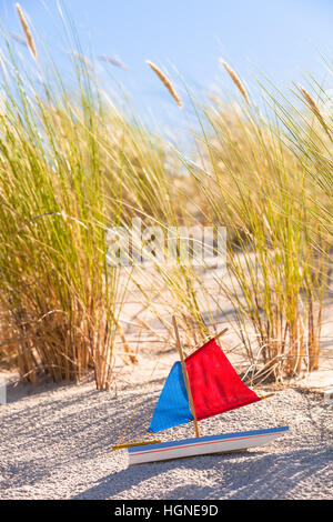 Coastal landscape with dune grass and old toy boat with red and blue colored sails in the sand Stock Photo