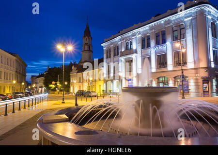 Vilnius, Lithuania - July 8, 2016: Gray Marble Fountain With Water Jets On Illuminated Rotuses Square. Steeple Of St. Nicholas Church Behind On Summer Stock Photo
