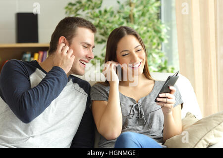 Happy couple listening music and sharing earphones sitting on a sofa in the living room at home Stock Photo