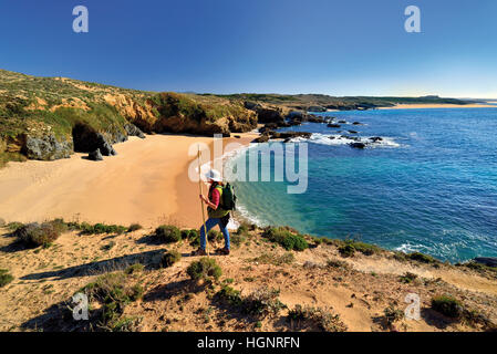 Woman with backpack and walking stick trekking along a sandy coastal trail along lonely beaches and a calm blue ocean Stock Photo