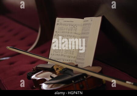 Violin with bow laying on dark red church pew with hymnal leaning against it and reflecting in wooden backrest Stock Photo