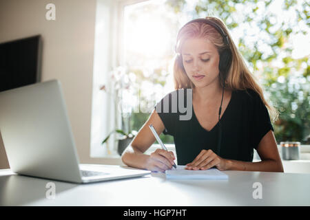 Indoor shot of woman taking notes in kitchen. Female wearing headphones and writing with laptop on table. Stock Photo