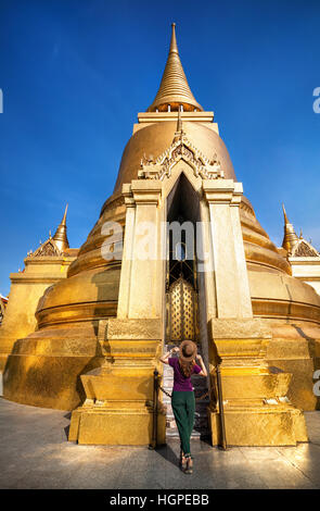 Woman tourist walking in the Temple of the Emerald Buddha near big Golden Stupa in Bangkok at sunset Stock Photo