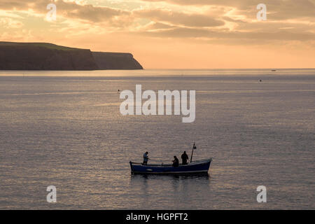 3 men, aboard a small boat, on a sea fishing trip at sunset - Whitby, with Sandsend beyond, North Yorkshire, England. Stock Photo