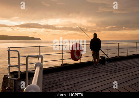 Scenic Whitby & silhouetted against dramatic, bright colourful sunset sky, man (angler) is sea fishing from West Pier - North Yorkshire, England, UK. Stock Photo