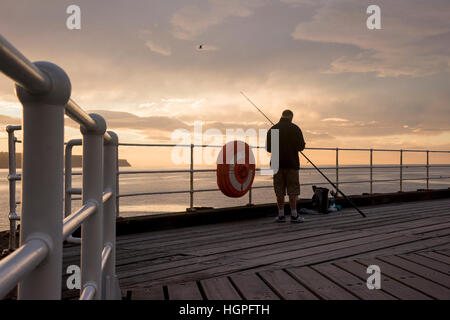 Scenic Whitby & silhouetted against dramatic, bright colourful sunset sky, man (angler) is sea fishing from West Pier - North Yorkshire, England, UK. Stock Photo