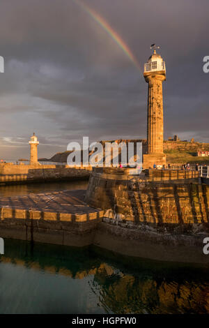 Rainbow and dark sky over sunlit lighthouses and harbour entrance, Abbey on the cliffs beyond - Whitby, North Yorkshire, GB. Stock Photo
