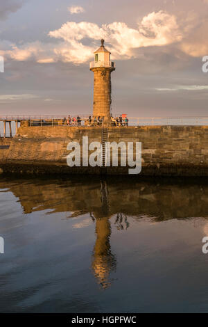 East Pier lighthouse, harbour entrance and people, lit by evening sun and reflected in the sea - Whitby, North Yorkshire, GB. Stock Photo
