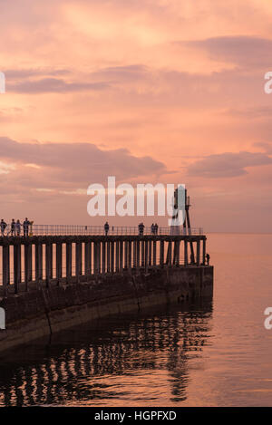 At sunset, people and pier are silhouetted against the dramatic, bright, red sky and sea - Whitby, North Yorkshire, England. Stock Photo