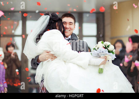 The groom carrying bride in his arms, the crowd throws petals and rice. Happy wedding. Stock Photo